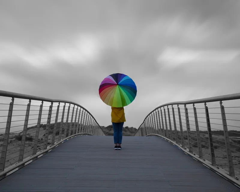 A woman walking away over a footbridge, holding a rainbow umbrella under a grey sky